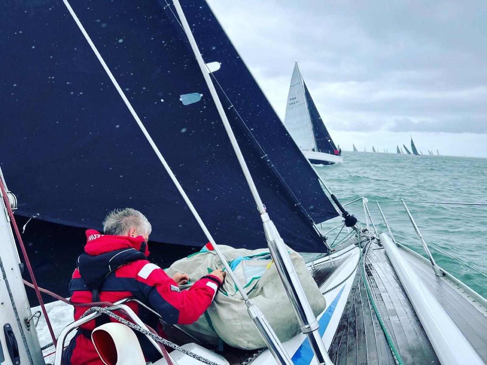 Crew working on SY Cormorant's foredeck mid-race, with the fleet astern